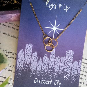 K&ouml;nnte beinhalten: Eine goldene Halskette mit drei ineinandergreifenden Kreisen h&auml;ngt an einer Karte mit violettem Hintergrund. Die Karte hat ein Stadtbild-Design und den Text "Light It Up" und "Crescent City".
