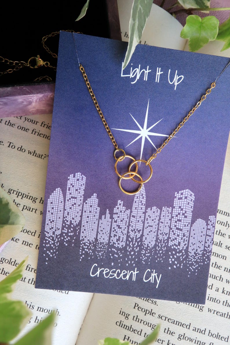 K&ouml;nnte beinhalten: Eine goldene Halskette mit drei ineinandergreifenden Kreisen h&auml;ngt an einer Karte mit violettem Hintergrund. Die Karte hat ein Stadtbild-Design und den Text "Light It Up" und "Crescent City".