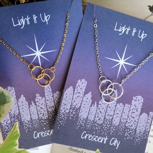 K&ouml;nnte beinhalten: Zwei Halsketten-Displays mit dem Text "Light It Up" und "Crescent City". Die Halsketten haben eine goldene und silberne Kette mit einem dreifachen Kreis-Anh&auml;nger.