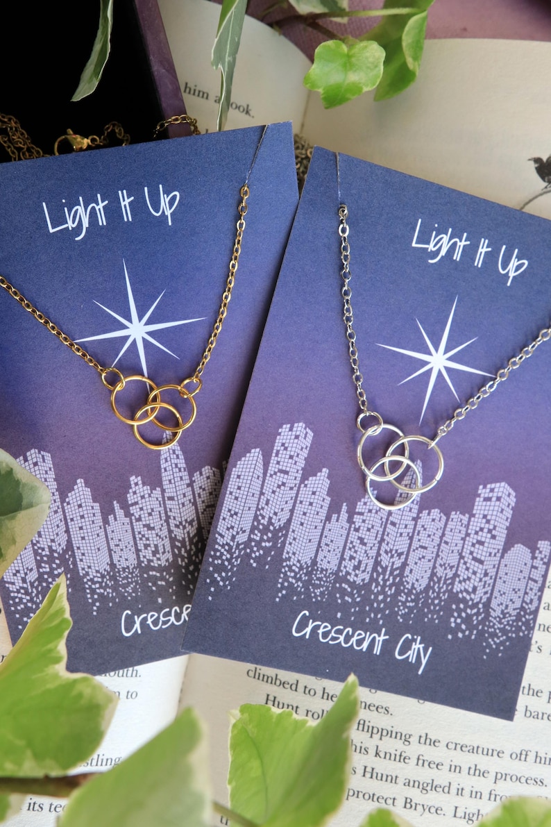 K&ouml;nnte beinhalten: Zwei Halsketten-Displays mit dem Text "Light It Up" und "Crescent City". Die Halsketten haben eine goldene und silberne Kette mit einem dreifachen Kreis-Anh&auml;nger.
