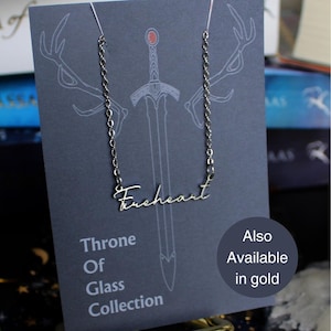 May include: A silver necklace featuring the word "Fireheart" in a cursive script. The necklace is presented on a grey card with a sword and antlers. The card also states "Throne of Glass Collection" and "Also Available in Gold".