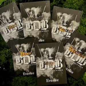 May include: A collection of gold and silver-toned nameplate necklaces on display. Each necklace features a different name or word, suspended from a delicate chain. The cards behind the necklaces have a sword graphic and the text "OFFICIALLY LICENSED".