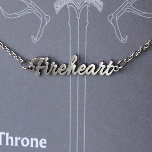 Puede incluir: Un collar plateado con la palabra "Fireheart" en cursiva. El collar está sobre un fondo gris con una espada y otros elementos de diseño. El texto "Throne of Glass" es visible en la parte inferior.