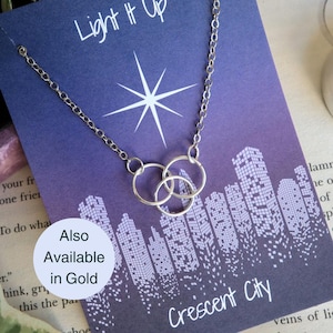 K&ouml;nnte beinhalten: Eine silberne Halskette mit drei ineinander verschlungenen Kreisen, die an einer Karte mit einem violetten Stadtbild im Hintergrund und dem Text "Light It Up Crescent City" h&auml;ngt. Die Karte sagt auch "Also Available in Gold".