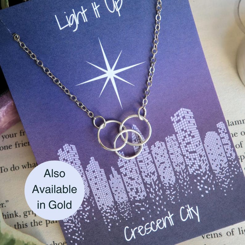 K&ouml;nnte beinhalten: Eine silberne Halskette mit drei ineinander verschlungenen Kreisen, die an einer Karte mit einem violetten Stadtbild im Hintergrund und dem Text "Light It Up Crescent City" h&auml;ngt. Die Karte sagt auch "Also Available in Gold".