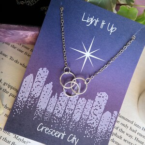 K&ouml;nnte beinhalten: Eine silberne Halskette mit drei ineinandergreifenden Kreisen h&auml;ngt an einer lila Karte mit einem wei&szlig;en Stern und dem Text "Light It Up". Die Karte zeigt auch ein Stadtbild mit dem Text "Crescent City".