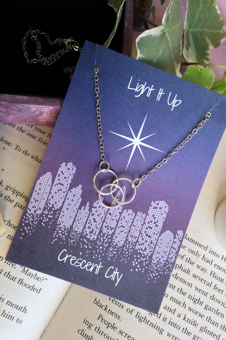 K&ouml;nnte beinhalten: Eine silberne Halskette mit drei ineinandergreifenden Kreisen h&auml;ngt an einer lila Karte mit einem wei&szlig;en Stern und dem Text "Light It Up". Die Karte zeigt auch ein Stadtbild mit dem Text "Crescent City".