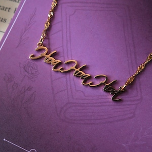 May include: A gold-coloured necklace spelling "Her Hero" in a cursive font. The necklace is presented on a purple background, with the brand name "Everlee Jew" visible. The chain has a delicate, twisted design.