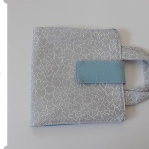 Könnte beinhalten: Eine graue Stofftasche für Bücher mit einem hellblauen Griff und einer hellblauen Klappe mit Klettverschluss. Die Tasche ist mit einem weißen Herzmuster bedeckt.