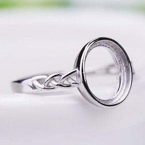 May include: A silver ring with a round, open setting for a stone or cameo. The ring band is a braided design.