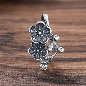 Puede incluir: Un colgante de plata con dos flores detalladas de cinco pétalos unidas a una rama texturizada con pequeños brotes. El metal tiene un acabado oxidado que resalta los intrincados patrones florales y la forma orgánica del tallo y las flores.