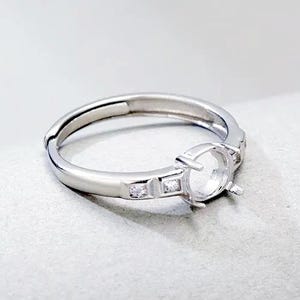 May include: A silver-toned ring featuring a central circular prong setting designed to hold a gemstone. The band includes two small, square-cut clear stones embedded on each side of the main setting, creating a polished and metallic appearance.