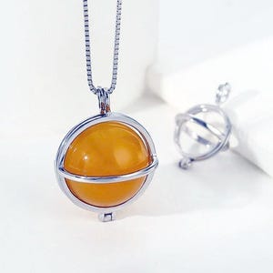 May include: A silver-toned necklace featuring a spherical pendant with a smooth, translucent orange stone encased in a metal cage frame. A second, empty silver-toned spherical cage pendant sits nearby. The chain is a thin, delicate box link style.