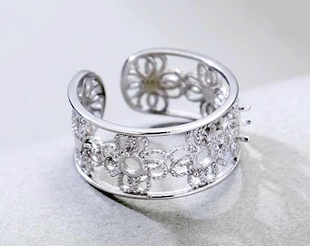 Könnte beinhalten: Ein silberfarbener offener Ring mit einem sich wiederholenden Blumenmuster entlang der Ringschiene. Das Design besteht aus miteinander verbundenen kreisförmigen Blumenmotiven, die mit kleinen, klaren, funkelnden Steinen besetzt sind, die das Licht einfangen.