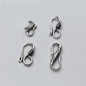 Può includere: Quattro chiusure per gioielli in metallo color argento disposte in una griglia. Ogni chiusura a gancio presenta un distinto motivo testurizzato o inciso sul braccio curvo. I componenti metallici hanno una finitura lucida con ossidazione scura.