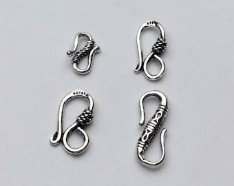 May include: Four silver-toned metal jewelry clasps arranged in a grid. Each hook-style clasp features a distinct textured or engraved pattern on the curved arm. The metal components have a polished finish with dark oxidation in the recessed areas.