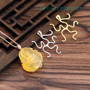 Peut inclure: Un pendentif Bouddha jaune translucide sur une chaîne en or rose, accompagné de deux breloques métalliques abstraites aux tons argent et or. Le texte ClareAccessories est visible dans la partie supérieure. Les objets sont disposés ensemble.