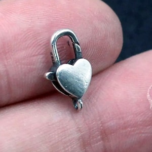 Op de afbeelding: Een kleine, zilverkleurige hartvormige sluiting. De sluiting heeft een scharnierende bovenkant en een kleine lus voor bevestiging. Het hart is gedetailleerd met een licht getextureerd oppervlak. De sluiting wordt in de hand van een persoon gehouden.