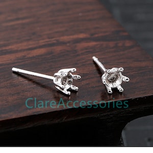 May include: Two silver earring settings with prongs for holding a gemstone. The settings are on a wooden surface.