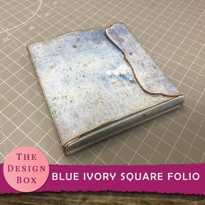 May include: A square folio with a blue and ivory lace design. The folio has a flap closure and gold accents. The text "BLUE IVORY SQUARE FOLIO" is visible on the bottom right of the image. The Design Box logo is on the bottom left.