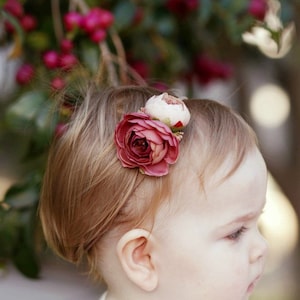 May include: A child's headband featuring a large burgundy rose and a smaller white flower. The headband is a thin, neutral colour, and the flowers are the focal point. The background is blurred with pink flowers.