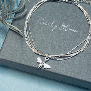 May include: A silver multi-strand bracelet with a small bee charm. The bracelet is on a grey background with the text "Ciely Bloom" printed on it.