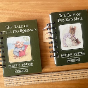 May include: Two small, spiral-bound books with green covers. One features a pig with a spyglass, the other a mouse. Both books are titled "The Tale of..." by Beatrix Potter. A ruler is at the bottom, and a tag hangs from one book.
