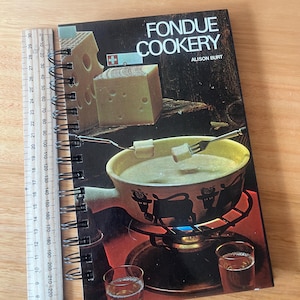 May include: A spiral-bound cookbook titled "Fondue Cookery" by Alison Burt. The cover features a fondue pot over a burner, cheese, and glasses. A ruler is placed next to the book, showing measurements in centimeters.