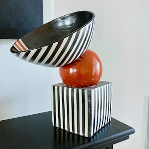 Op de afbeelding: Een zwarte en witte gestreepte keramische kom staat op een rode bol, die op een zwarte en witte gestreepte kubus is gebalanceerd. De kom heeft een rode streep rond de rand.
