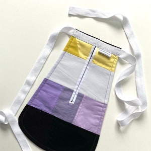 May include: A black, purple, white and yellow fabric apron with a white zipper and white ties. The apron has a label that says "Handmade with Love".