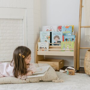May include: A light-colored wooden bookshelf holds several children's books with colorful covers, including titles like Albert Einstein, Dream Big Little One, and Llamaste & Friends, displayed in a room with a white fireplace and a wooden ladder.