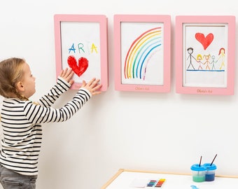 May include: Three rectangular pink wall-mounted frames for displaying children's artwork. Each frame features a light wood finish and a personalized name engraved at the bottom. The frames contain drawings of a heart, a rainbow, and a family portrait.