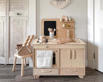 May include: A wooden play kitchen with a white towel hanging from the oven door, a basket of bread rolls, and a sign that reads "PLAY is the WORK of the CHILD".