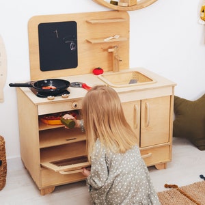 May include: A child plays with a wooden toy kitchen set. The kitchen features a stove with an oven, a sink, and a chalkboard. The child is wearing a green and white polka dot dress and is opening the oven door.