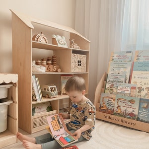 Könnte beinhalten: Ein hölzernes, hausförmiges Bücherregal mit Kinderbüchern und Spielzeug. Ein Kind sitzt auf dem Boden und liest ein Buch. Das Bücherregal hat mehrere Regale mit verschiedenen Gegenständen.