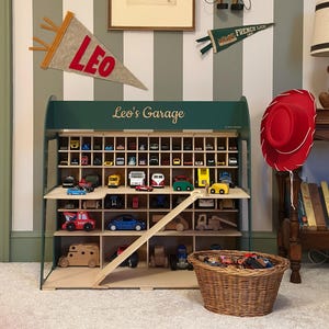 K&ouml;nnte beinhalten: Ein Holzregal f&uuml;r Spielzeugautos mit der Aufschrift "Leo's Garage", gef&uuml;llt mit Miniatur-Spielzeugautos. Ein roter Cowboyhut h&auml;ngt in der N&auml;he. Ein gerahmtes Pferdeportr&auml;t und eine Pinocchio-Puppe sind ebenfalls zu sehen.