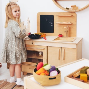 May include: A young girl plays with a wooden toy kitchen set. The kitchen features a stove, oven, sink, and cabinets. The girl is wearing a light green dress with white polka dots. There are toy vegetables in a basket on the counter.