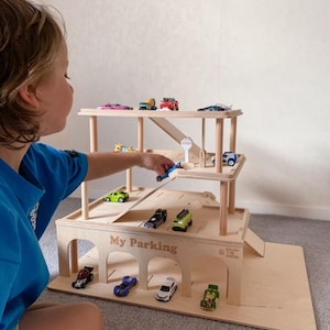 K&ouml;nnte beinhalten: Ein h&ouml;lzernes Spielzeug-Parkhaus mit mehreren Ebenen und Rampen. Das Parkhaus ist mit bunten Spielzeugautos gef&uuml;llt. Der Text "My Parking" ist auf der Vorderseite zu sehen. Das Spielzeug ist aus hellem Holz gefertigt.