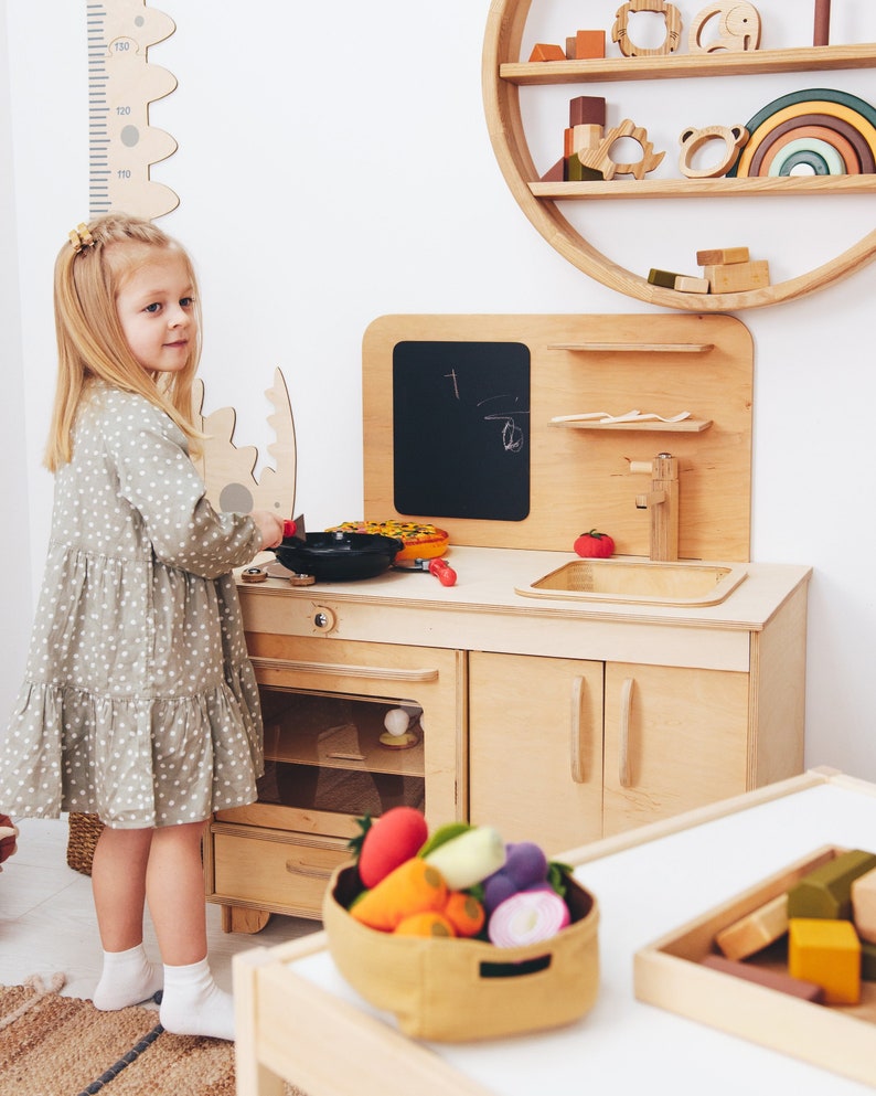 May include: A young girl plays with a wooden toy kitchen set. The kitchen features a stove, oven, sink, and cabinets. The girl is wearing a light green dress with white polka dots. There are toy vegetables in a basket on the counter.