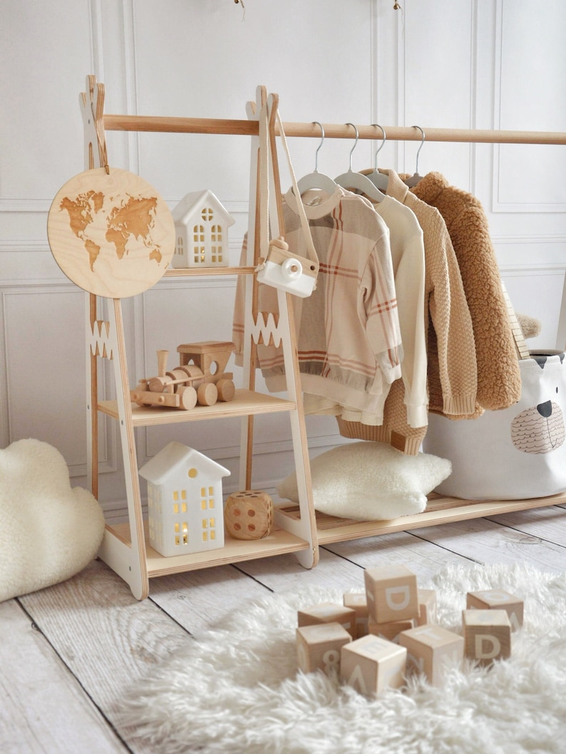 Puede incluir: Un perchero de ropa para ni&ntilde;os de madera con acabado blanco y madera. El perchero tiene una barra para colgar, dos estantes y un peque&ntilde;o tren de madera en el estante inferior. Tambi&eacute;n hay dos casas de cer&aacute;mica blanca y un mapa del mundo de madera en el perchero. El perchero est&aacute; sobre una alfombra blanca y esponjosa con bloques de letras de madera esparcidos alrededor.