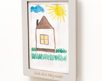 May include: A light gray rectangular picture frame containing a child's drawing of a house, a yellow sun, and a blue cloud. The frame features the gold text "Look what Abby made!" printed on the bottom border, displaying the artwork clearly.