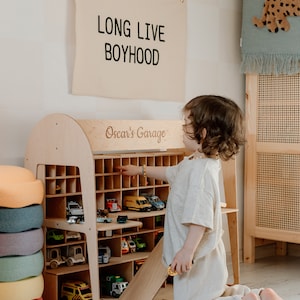 K&ouml;nnte beinhalten: Ein Holzspielzeug-Autogarage mit mehreren F&auml;chern, gef&uuml;llt mit verschiedenen Spielzeugautos. Das Schild oben lautet "LONG LIVE BOYHOOD". Die Garage ist mit "Oscar's Garage" beschriftet. Ein Kind interagiert mit den Spielzeugautos.