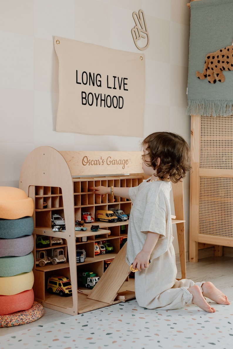 K&ouml;nnte beinhalten: Ein Holzspielzeug-Autogarage mit mehreren F&auml;chern, gef&uuml;llt mit verschiedenen Spielzeugautos. Das Schild oben lautet "LONG LIVE BOYHOOD". Die Garage ist mit "Oscar's Garage" beschriftet. Ein Kind interagiert mit den Spielzeugautos.