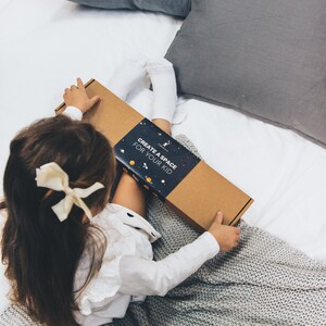 以下が含まれることがあります： CREATE A SPACE FOR YOUR KIDという白い文字が書かれた濃い青色のスリーブが付いた長方形の段ボール箱。表面には惑星、星、人工衛星の小さなオレンジ色と白色のイラストが描かれており、白い背景の上に置かれています。