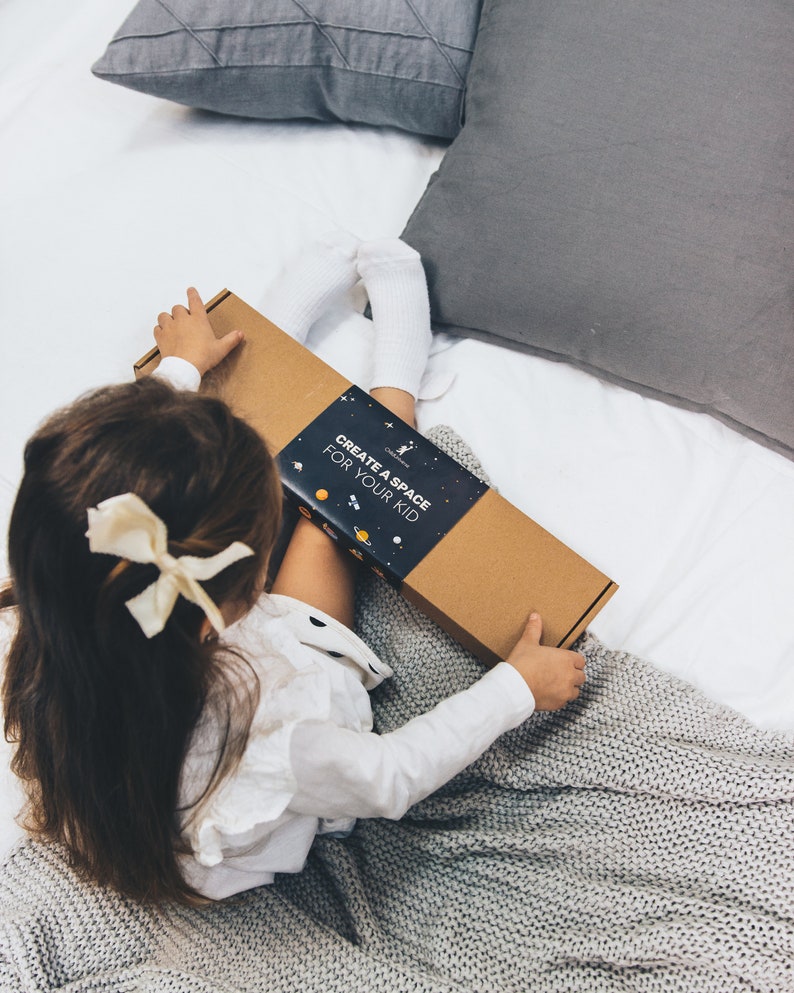以下が含まれることがあります： CREATE A SPACE FOR YOUR KIDという白い文字が書かれた濃い青色のスリーブが付いた長方形の段ボール箱。表面には惑星、星、人工衛星の小さなオレンジ色と白色のイラストが描かれており、白い背景の上に置かれています。