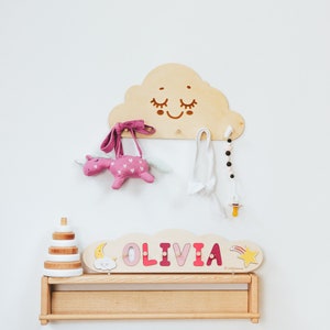 May include: Wooden wall-mounted cloud-shaped rack with a smiling face, featuring a pink unicorn stuffed animal, a pink headband, a white headband, a beaded necklace, and a yellow pacifier. Below the rack is a wooden shelf with a stack of wooden rings and a personalized name sign that reads "OLIVIA".