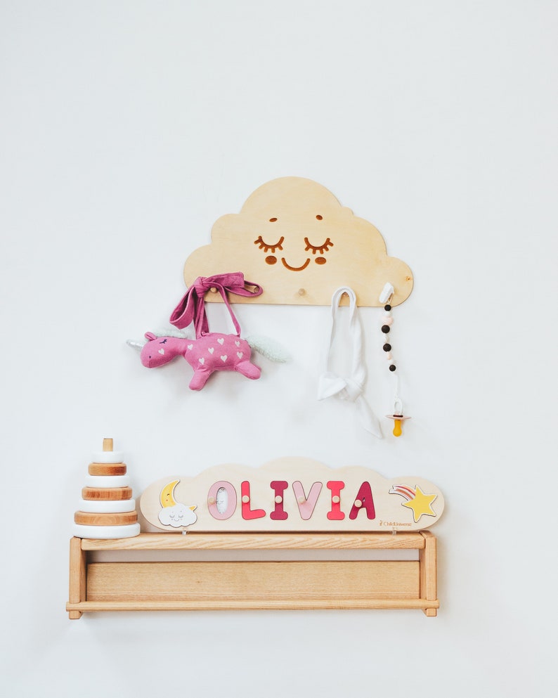 May include: Wooden wall-mounted cloud-shaped rack with a smiling face, featuring a pink unicorn stuffed animal, a pink headband, a white headband, a beaded necklace, and a yellow pacifier. Below the rack is a wooden shelf with a stack of wooden rings and a personalized name sign that reads "OLIVIA".