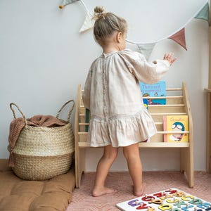 May include: A light beige long-sleeved dress with a button-down back, a wooden bookshelf holding children's books, a woven seagrass basket with a brown cloth, and a white wooden alphabet puzzle board with colorful letters.