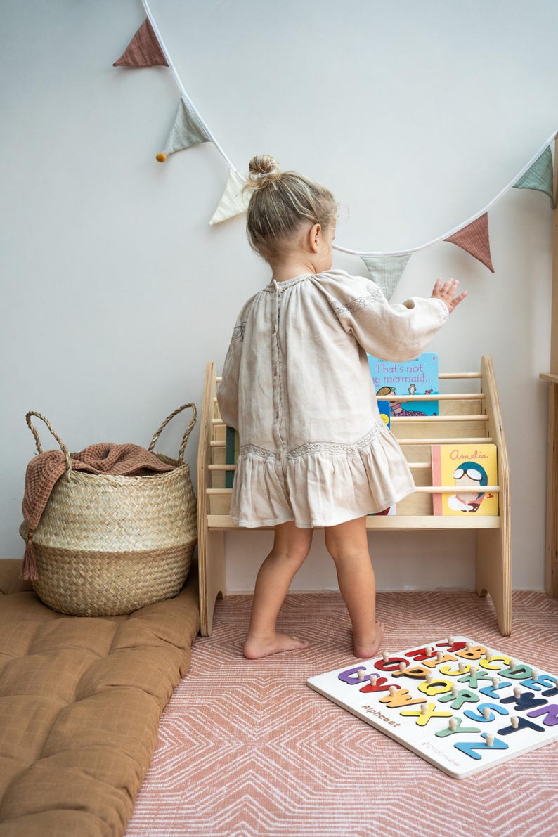 May include: A light beige long-sleeved dress with a button-down back, a wooden bookshelf holding children's books, a woven seagrass basket with a brown cloth, and a white wooden alphabet puzzle board with colorful letters.