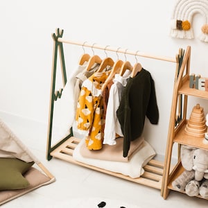 Puede incluir: Un perchero de madera con un dise&ntilde;o verde y marr&oacute;n, que incluye una barra para colgar y un estante. El perchero est&aacute; decorado con una variedad de ropa para ni&ntilde;os, incluyendo una camisa con estampado amarillo y negro, una camisa verde y una camisa blanca.